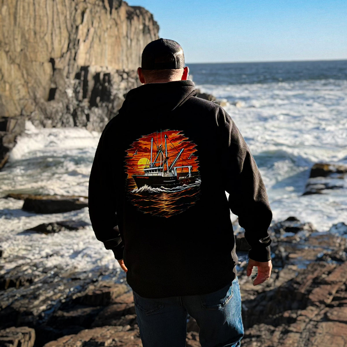 A man stands on a rocky coastline facing the ocean, wearing a black hoodie with a large back graphic of a commercial fishing trawler boat on orange and red waves, and blue jeans.